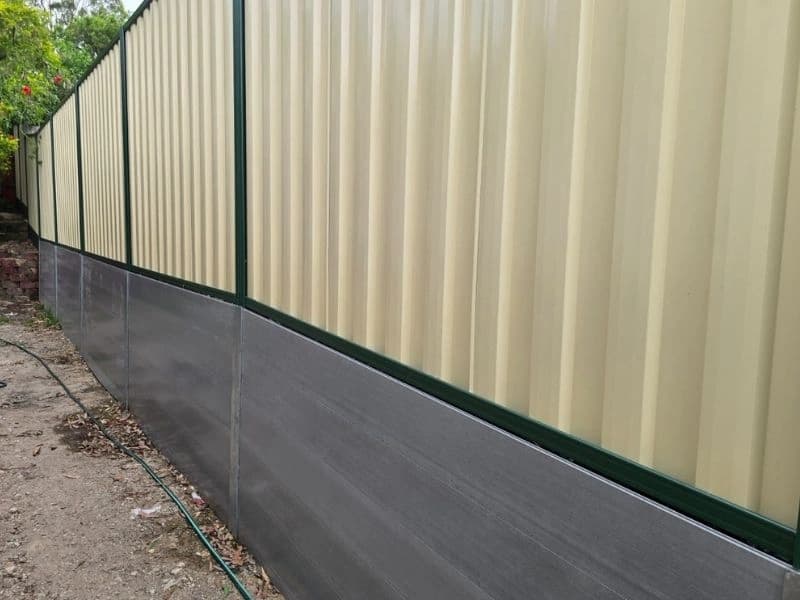 Cream Colorbond fence with green rails above a grey concrete sleeper retaining wall